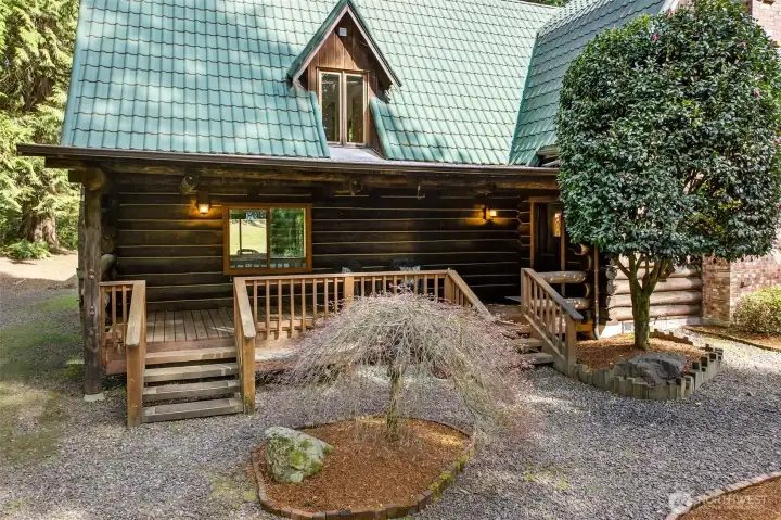 The main entry porch with log-post framing and a dormer window above sets an inviting tone, complemented by thoughtfully curated plantings and a gravel motor court that adds to the property's natural, private character.