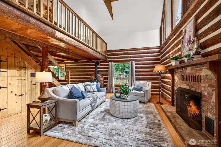 The great room rises dramatically with soaring vaulted ceilings, a floor-to-ceiling brick fireplace, and a loft overlook above — a truly one-of-a-kind living space where the craftsmanship of hand-laid log walls sets the tone for the entire home.