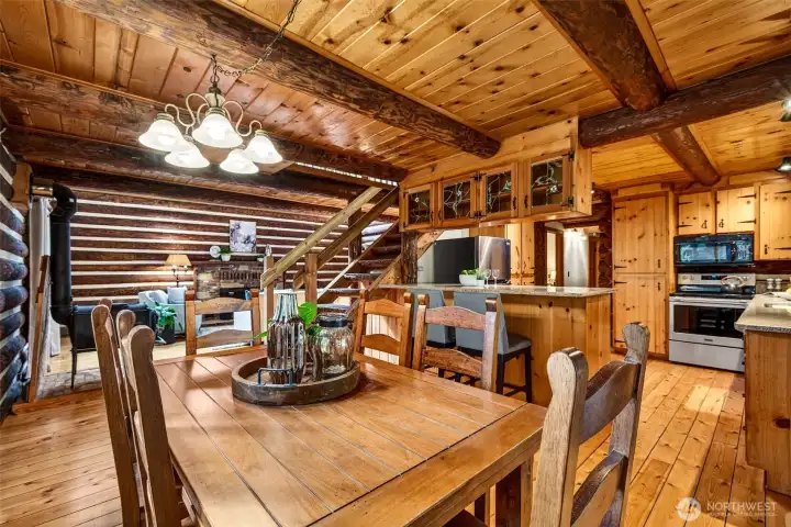 The open flow between the dining area, kitchen, and great room makes everyday living and entertaining effortless — hand-hewn log walls, a knotty pine ceiling with exposed timber beams, and warm hardwood floors connect every space with authentic cabin character.