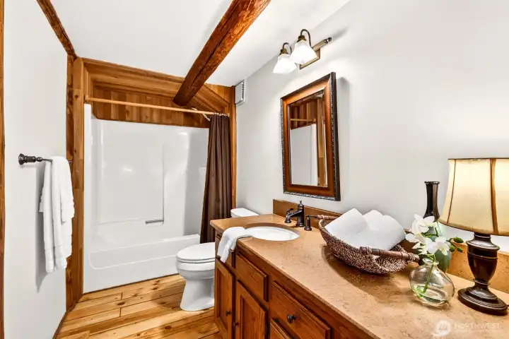 The updated full hall bath balances cabin warmth with clean, modern comfort — wood-trimmed accents, a tub and shower combination, and a well-appointed vanity with stone countertop deliver a polished and functional space for guests and household alike.