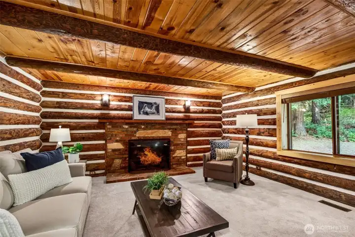 The second fireplace in the main-level living area anchors the room with warmth and character — rich log wall construction on all sides and tree canopy views through the window create a cozy, immersive atmosphere that feels like a permanent retreat.