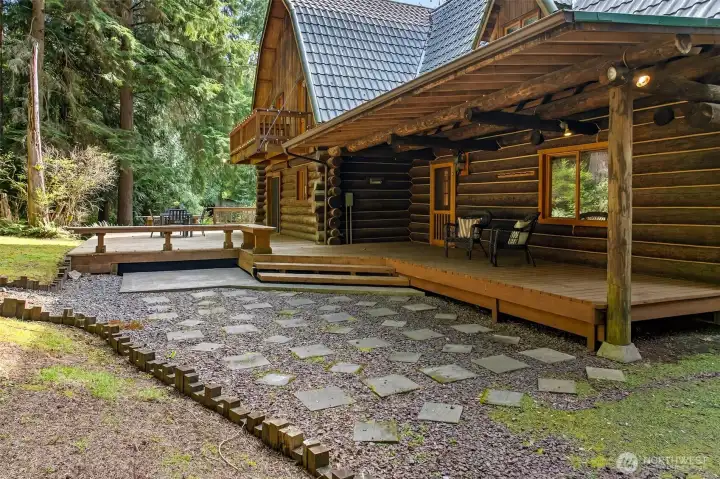 The back of the home reveals an impressive multi-level outdoor living layout — a covered porch, an expansive deck, and a stepping-stone pathway through the landscaped grounds invite year-round enjoyment of the property's spectacular natural setting.