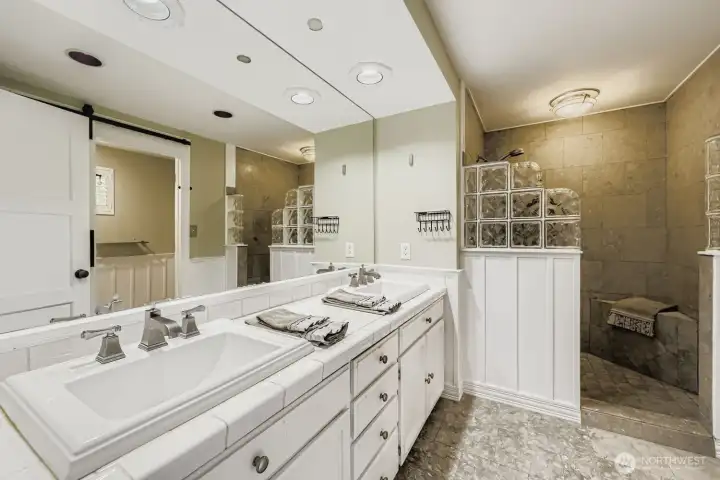 The well-designed Primary bath boasts a large walk-in tiled shower with glass blocks, dual sinks, excellent lighting, abundant cabinetry, tile countertops and flooring.