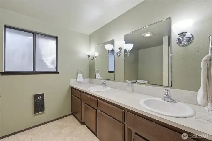 En suite primary bathroom features double sinks, privacy window for natural light, and ample space. Opposite the long vanity and double mirrors are the shower and commode.