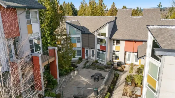 This inviting townhome community courtyard features modern multi-colored units with sloped roofs and large windows, surrounded by lush evergreens, a central stone patio with seating, low-maintenance landscaping, and private balconies overlooking the serene wooded setting.