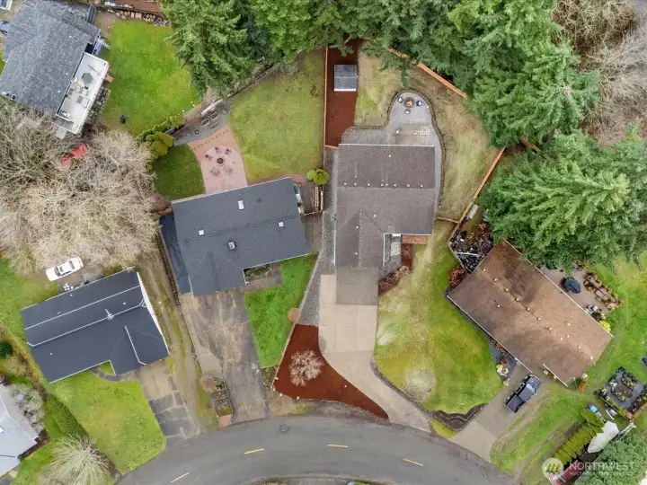 Aerial view showcasing the property's generous lot, curved driveway, private fenced backyard, firepit area, and surrounding mature trees in a quiet residential setting