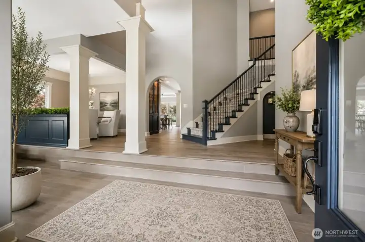 (virtually staged)   Welcoming front entry showcasing a grand foyer with tall ceilings, architectural columns, and a statement staircase. Wide-plank flooring and an open sightline into the main living areas create a refined first impression and seamless flow throughout the main level.