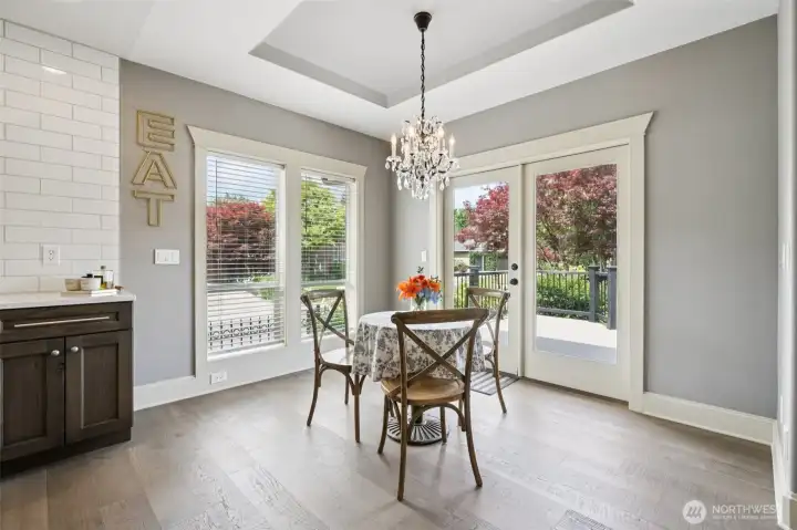 Light-filled dining area featuring tray ceiling detail, statement chandelier, and direct access to the outdoor deck through French doors. Large windows provide natural light and views of the landscaped yard, creating a seamless flow between indoor dining and outdoor living.