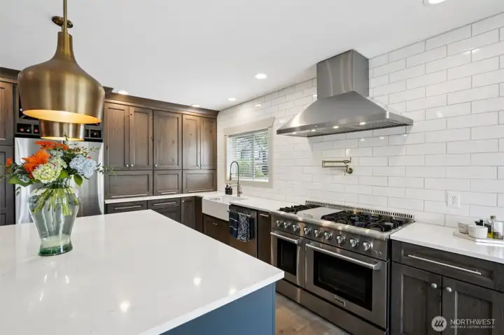 Well-appointed gourmet kitchen featuring custom appliance cabinetry, expansive quartz countertops, and a professional-grade double oven range with six burners and center griddle. Additional highlights include a statement vent hood, farmhouse sink, generous island seating, and thoughtful storage throughout, creating a highly functional space designed for both everyday use and entertaining.