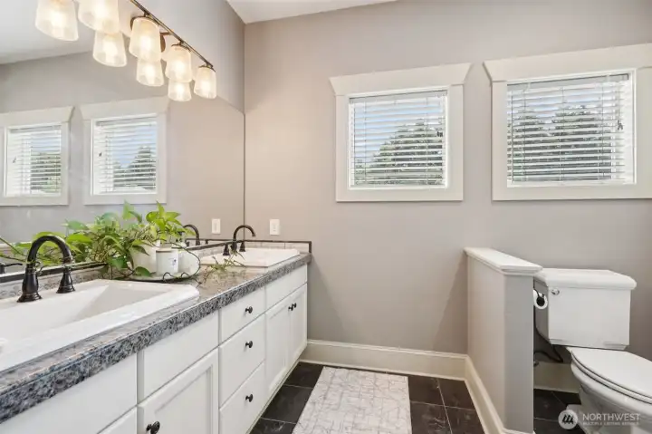 Well-appointed primary bathroom featuring a double vanity with granite countertops, ample cabinetry, and updated lighting. Large windows provide natural light while maintaining privacy, creating a bright and functional space designed for everyday comfort.