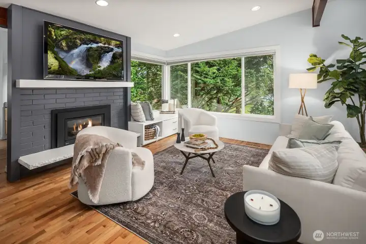 The bright and inviting living room features rich hardwood floors, a cozy fireplace, and an abundance of natural light pouring in through large windows, creating a warm and stylish space for everyday living and entertaining.