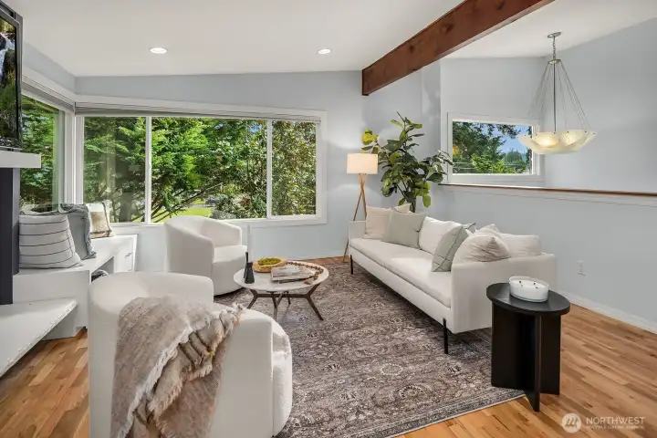 Soaring vaulted ceilings with elegant beam detailing, gleaming hardwood floors, and walls of windows framing lush green views give this living room a bright, airy, and sophisticated feel.