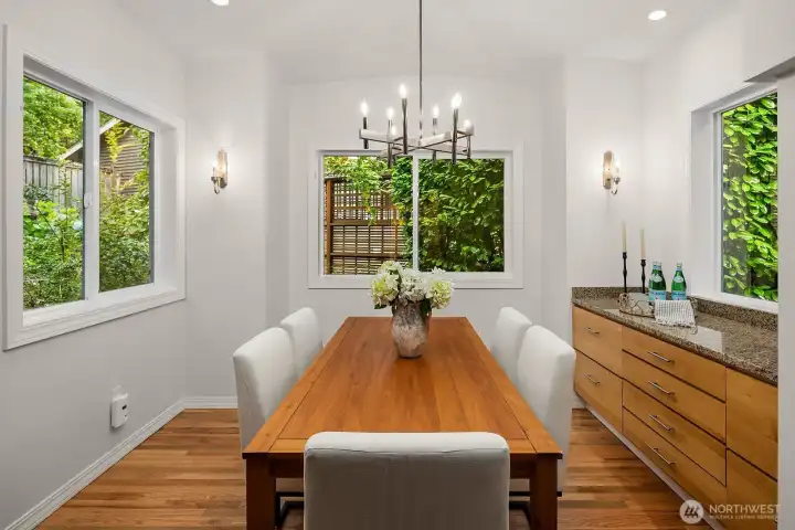 The elegant formal dining room impresses with a statement chandelier, beautiful hardwood floors, and walls of windows on two sides flooding the space with natural light and lush garden views, perfect for hosting memorable gatherings.