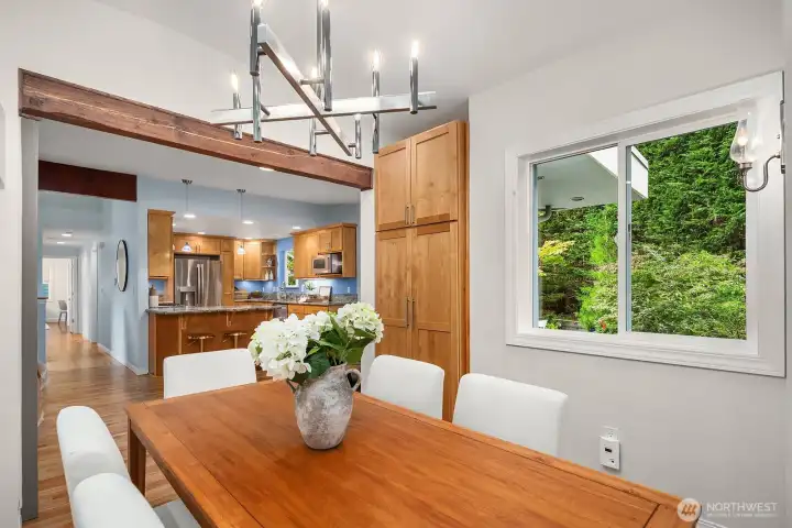 A stunning linear chandelier anchors this bright dining room, which flows effortlessly into the chef's kitchen beyond, with warm wood accents and lush garden views creating a naturally inviting atmosphere throughout.