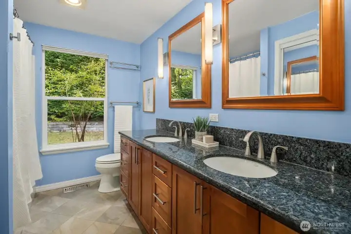 Flooded with natural light from a large window, this well-designed primary bath showcases dual granite vanities, warm custom cabinetry, and a clean, bright finish, a functional and elegant everyday retreat.