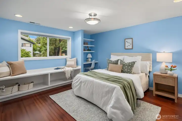 This generously sized lower level bedroom features warm hardwood floors, a large window with built-in bench seating, and custom shelving,  a thoughtfully designed and welcoming space for family or guests.