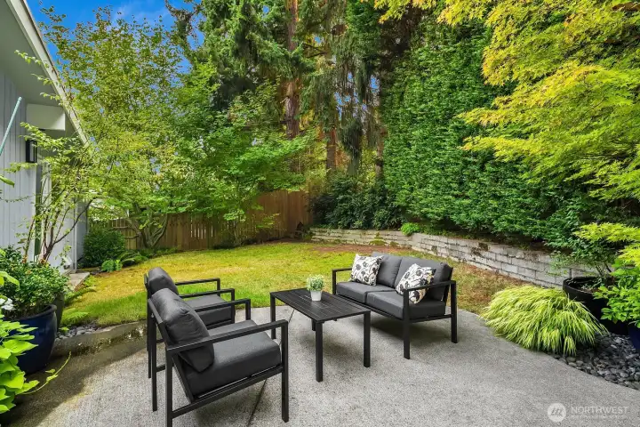 A beautifully landscaped and fully fenced backyard surrounds this inviting patio seating area with lush greenery on all sides, offering a peaceful and private outdoor sanctuary ideal for relaxing or entertaining guests.
