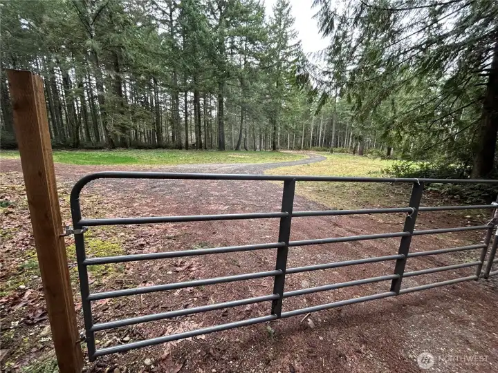 A gated entry with steel cattle-panel gate and wood post framing welcomes you onto the property. The gravel drive beyond curves gracefully through the trees, offering privacy and a sense of arrival.