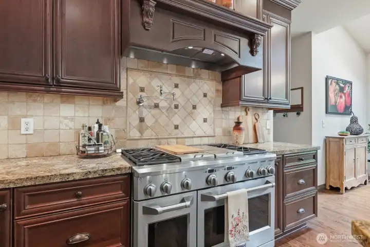 The professional-grade gas range features a griddle and grill, complemented by dual ovens, a pot filler, and a commercial-style hood. A full tile backsplash with designer accent details enhances the kitchen’s timeless appeal.