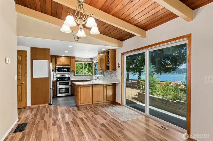 Open kitchen and dining area featuring vaulted wood ceilings, exposed beams, stainless steel appliances, and an oversized sliding glass door framing the lush outdoor setting and bay views. Open kitchen and dining area featuring vaulted wood ceilings, exposed beams, stainless steel appliances, and an oversized sliding glass door framing the lush outdoor setting and bay views.