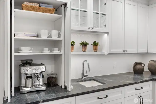 This elegant coffee bar is tucked behind custom cabinet doors for a clean, seamless look when not in use. Outfitted with a Breville espresso machine, open shelving holds mugs, dishes, and entertaining essentials within easy reach.
