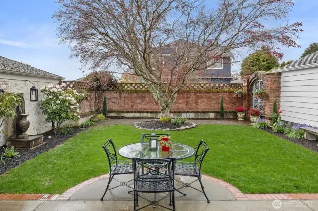 The patio flows effortlessly into a lush lawn bordered by manicured garden beds, mature rhododendrons, and thoughtfully placed perennials.