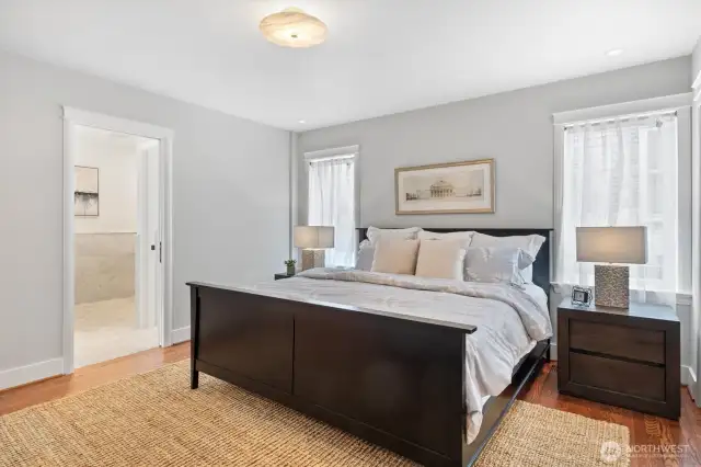 This serene main floor primary suite features a soothing neutral palette, hardwood floors, and abundant natural light filtered through dual windows. A sculptural alabaster flush-mount light adds a touch of elegance overhead, while wide baseboards and crisp millwork offer a tailored finish.    Generous closet space is tucked behind classic paneled sliding doors, and an en suite bath—accessible through a pocket door—enhances privacy and convenience. Designed for comfort and functionality, this suite provides a peaceful retreat just steps from the heart of the home.