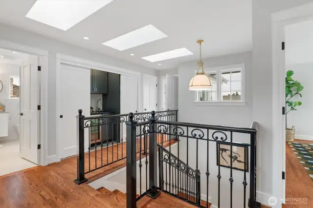 The upper landing is filled with natural light from a series of Velux skylights, one of which features automatic moisture-sensing closure for added peace of mind. Custom wrought-iron railings lend architectural character and enhance the sense of openness, while warm hardwood floors and classic millwork ground the space in timeless appeal. Just to the right of the wet bar, a front-load stacked Bosch ventless condenser washer and dryer adds discreet convenience.