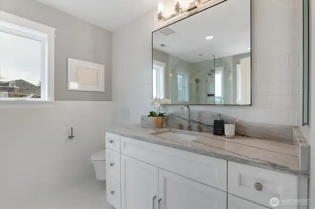 This thoughtfully designed en-suite features dual closets and elegant White Pearl Quartzite countertops with an ogee edge. An undermounted sink is paired with Top Knobs satin nickel hardware, while a 42"x36" floating mirror from Rejuvenation and a Robern medicine cabinet add both style and storage.  The glass-enclosed shower includes a bench and is outfitted with Kohler’s DTV digital thermostatic system, offering a programmable experience with both showerhead and hand shower in polished nickel. A wall-mounted toilet and heated subway tile floors with wainscoting complete the space—delivering comfort, craftsmanship, and timeless appeal.
