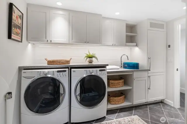 This beautifully designed laundry and utility area is as functional as it is stylish, featuring Kenmore Elite XL front-load washer and dryer, an integrated sink with Grohe polished chrome faucet, and generous counter space with under-cabinet lighting. A built-in Liebherr refrigerator to the right of the sink adds convenience and versatility.
