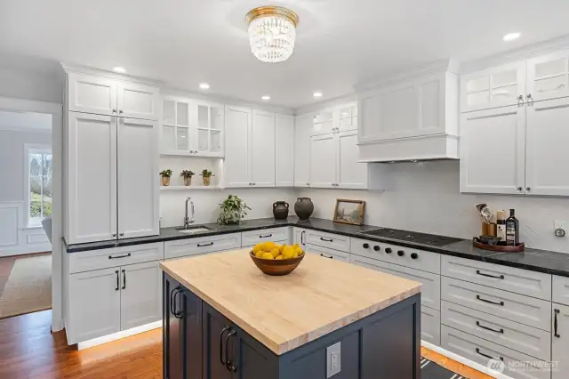 This expertly crafted kitchen pairs timeless materials with modern functionality. Water Street Brass oil-rubbed bronze pulls and crystal knobs lend refined elegance to the custom cabinetry. A Sonoma Herringbone matte white backsplash, paired with durable Mapei Kerapoxy grout, adds both texture and practicality.    Custom undermount and toe-kick lighting enhance the workspace and create an inviting ambiance, while a warm butcher block island anchors the room—perfect for casual dining, prep, or gathering with guests.