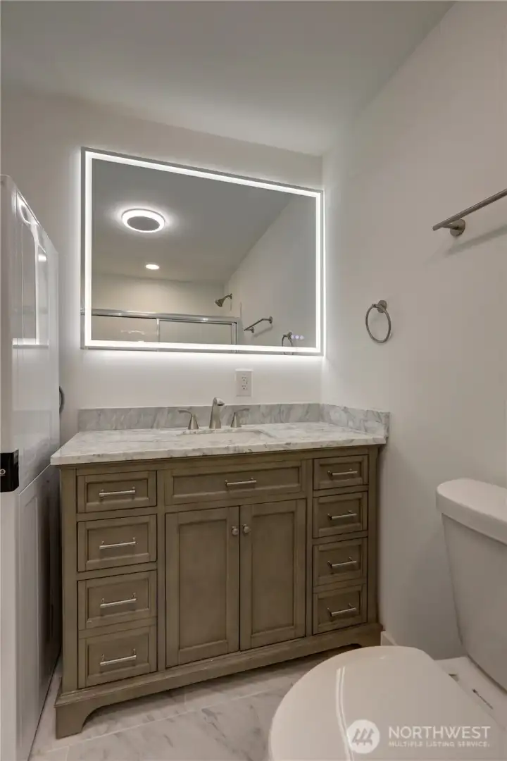 A modern minimalist bathroom featuring a marble countertop vanity, illuminated mirror, and sleek neutral-toned finishes.