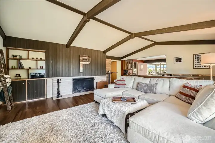Mid-century warmth meets modern comfort in this inviting living room featuring vaulted ceilings with exposed beams, rich wood-paneled accents, and a classic wood-burning fireplace. Built-in shelving adds character and functionality, while the open flow to the dining and kitchen areas creates the perfect setting for entertaining or relaxing in style. A cozy, elevated vibe that feels like home the moment you walk in.