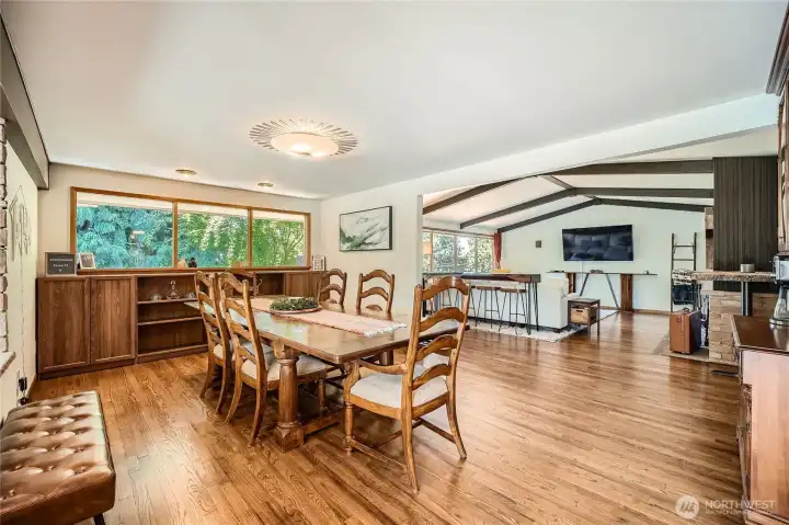 Beautifully connected and full of natural light, this spacious dining area flows seamlessly into the open-concept living room—creating the perfect layout for gatherings and everyday living. Large picture windows frame lush, tree-lined views, while rich hardwood floors and exposed beam ceilings elevate the mid-century charm. A statement light fixture adds elegance above the dining table, and the open sightlines make entertaining effortless from room to room.