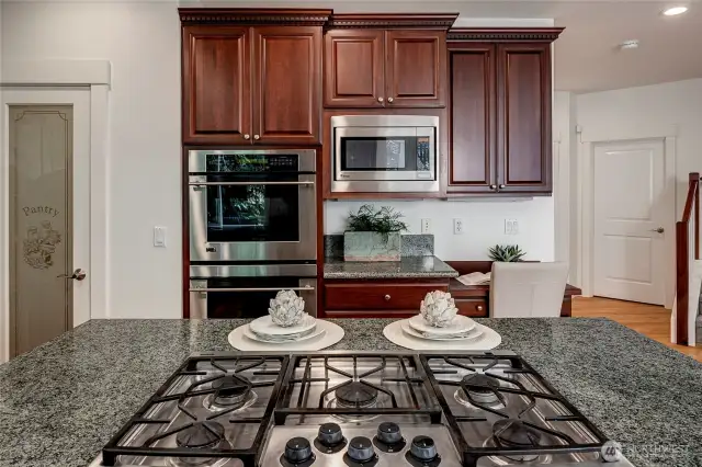 This view highlights the kitchen’s functional elegance with a sleek gas cooktop island, double wall ovens, built-in microwave, and a charming glass pantry door.