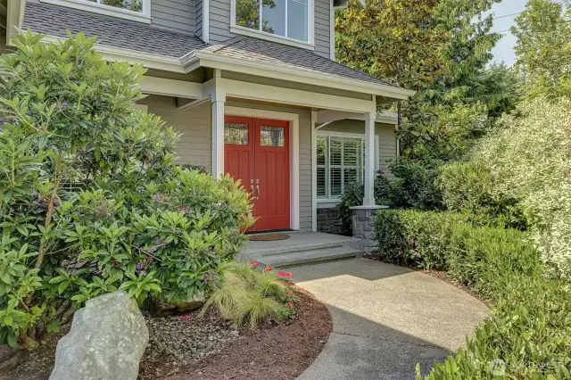A charming curved walkway guides you to a welcoming front porch framed by classic columns and lush, mature landscaping. The bold red double doors, accented with transom windows, make a memorable first impression and offer a pop of color against the home’s neutral siding.