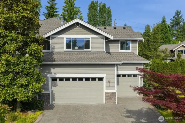 This side of the home showcases a spacious 3-car garage with two distinct doors—one double and one single—framed by crisp white trim and topped with transom-style windows that add architectural interest and natural light. The garage exterior features a combination of horizontal siding and stone accents at the base. The driveway is wide and cleanly finished, offering ample space for parking and easy access.