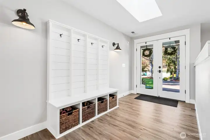 Right inside the door, this entry sets the tone for how the home functions. Double glass doors open fully, which makes everything from move-in day to summer gatherings feel effortless. Natural light from the skylight keeps the space bright, while the built-in coat and gear station keeps things organized without sacrificing style. Shiplap backing, hooks for everyday items, a bench for taking shoes on and off, and basket storage below all work together to keep clutter in check. The lighting has a modern edge with clean-lined sconces, and the flooring is a warm wood-look luxury vinyl plank that stands up well to traffic and weather. Soft gray walls and wide white baseboards give the whole space a calm, updated feel that flows nicely into the rest of the home.