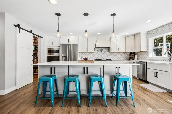 The island takes center stage with bright white quartz countertops that feel clean, polished, and ready for everything from meal prep to gathering. Above, three clear globe pendants add a contemporary touch without blocking sightlines, keeping the whole space feeling open and airy. The white tile backsplash runs all the way to the ceiling, which gives the kitchen a finished, elevated look that doesn’t cut corners. Shaker cabinets with sleek black hardware keep things crisp, and that barn door to the walk-in pantry adds just enough personality to stand out. The farmhouse-style apron sink sits perfectly in front of a large window overlooking the backyard, and sleek stainless steel appliances round everything out.