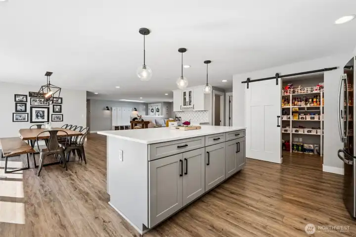 From the kitchen island you can see straight through to the dining area and back toward the entry, which gives the whole main space that open, connected feel buyers are always hoping for. The oversized island anchors the room and offers plenty of workspace and seating, while the wide plank luxury vinyl flooring runs seamlessly throughout, keeping everything cohesive and easy to maintain. You get a better look into the walk-in pantry here too, with its organized shelving that actually makes sense for real life. Recessed lighting carries across the ceiling, keeping the space bright and functional day to night. And tucked into the island, just out of full view, is a built-in beverage zone with a wine glass holder and beverage fridge, one of those features you’ll use more than you think.