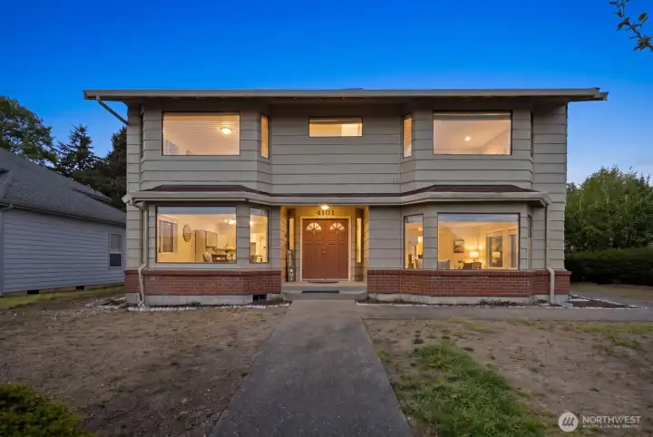 Front exterior at dusk with a covered entry, double front doors, and large windows showing the home lit from within