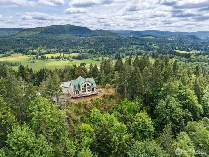 This aerial really captures the incredible sense of permanence & privacy this property offers b/c tucked into the surrounding forest & anchored directly into bedrock, this home was built w/serious long-term intention. The foundation itself was additionally reinforced & pinned directly into the rock below, creating exceptional structural stability on an already incredibly solid site, which honestly feels very on-brand for a property that approaches everything with a little extra “overbuild it & sleep peacefully forever” energy. Surrounding the homesite you can see the beautiful wooded acreage stretching outward in every direction creating that rare sense of total separation & quiet while still keeping the property practical & accessible. Both the home & the massive shop feature durable metal roofing adding long-term beauty, low-maintenance performance & that satisfying “this place was built to last” feeling throughout the entire compound. There is also a propane tank for future projects