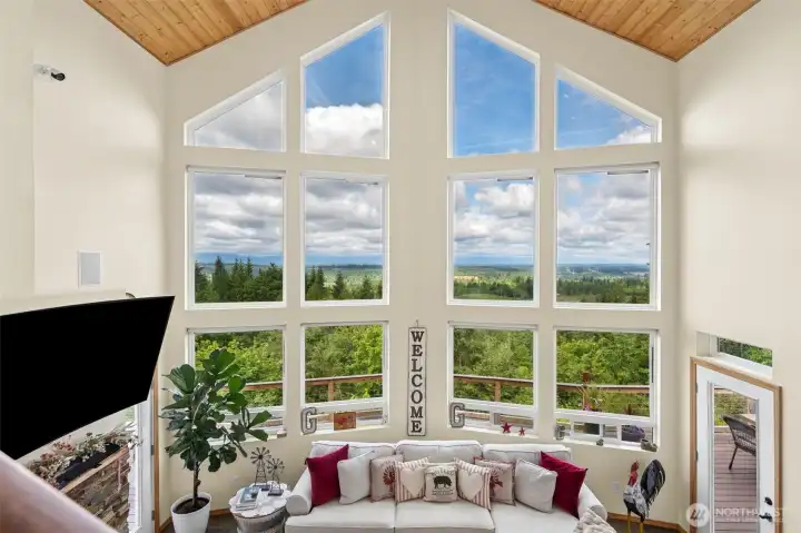 Standing at the top of the staircase, the scale of this home and the sheer magnitude of the views really hits you all at once because the massive vaulted showcase windows completely frame the Olympic Mountain range and surrounding territorial views like living artwork changing by the hour. This is not some tiny peekaboo mountain glimpse you have to lean sideways to find between rooftops either, this is full dramatic Pacific Northwest scenery on display from one of the best vantage points in the entire house. From up here you can also really appreciate how the soaring ceilings, wood-plank details and open sightlines all work together to create a true lodge-style atmosphere while still feeling bright and welcoming instead of dark or heavy, and just below, the glass door beside the fireplace leads directly out to the massive deck where stairs continue down toward the hot tub area.