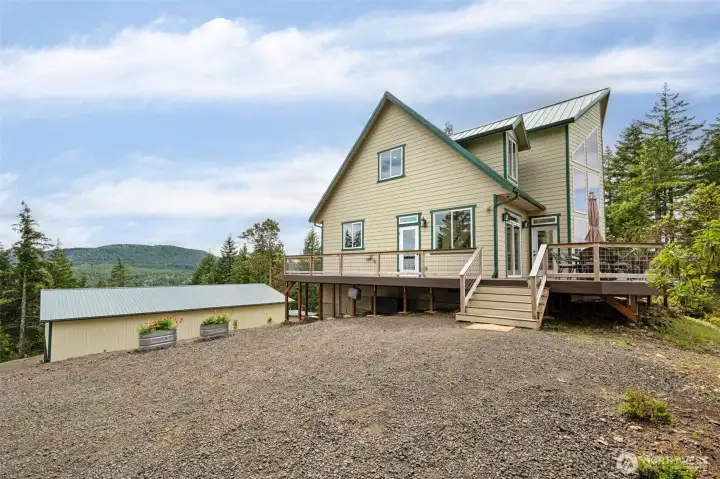 One of the underrated luxuries of this property is how incredibly easy and welcoming the arrival experience feels because unlike some rural mountain homes where guests need a map, a flashlight and emotional support to figure out where to park, this setup is wonderfully open, accessible and practical. The large gravel parking area offers abundant room for friends, family, recreational vehicles and easy turnaround space while the blooming planters add just enough softness and color to keep the approach feeling warm and inviting instead of overly utilitarian. The extra-wide staircase leading up toward the massive deck and main living areas also makes the transition to the home feel substantial and welcoming, and from this angle you can really appreciate how thoughtfully the property balances rugged mountain privacy with comfortable everyday usability.
