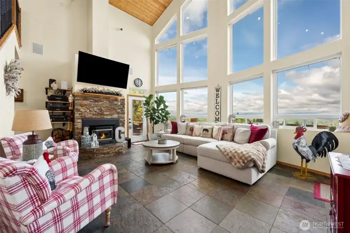 This is the room people will remember long after they leave the property. The massive 2-story vaulted great room feels almost cinematic w/its soaring wood-plank ceilings & towering showcase windows stretching across the entire wall, framing breathtaking Olympic Mts views like living artwork. Somebody designing this home clearly understood exactly what they were doing b/c the operable sections of the lower windows were thoughtfully positioned down at the base, allowing uninterrupted sightlines instead of chopping up the scenery w/bulky framing right at eye level. The result is an incredible full-sweep mt view that completely commands the room. A handsome stacked-stone fireplace w/gas insert grounds the space w/warmth & texture while the mounted TV remains w/the home & integrated speaker systems run throughout both the interior & exterior spaces w/individual rm controls for easy entertaining. Another glass door w/transom window above leads directly onto the huge 957-sf deck outside.