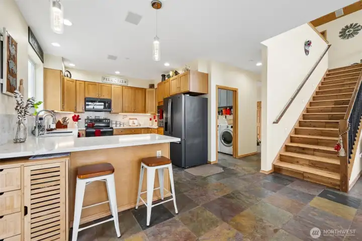 One of the things this home does exceptionally well is create separation w/o sacrificing openness & this view captures that perfectly. From the kitchen you can see how the layout naturally flows toward the foyer, laundry room & staircase while still keeping the kitchen itself slightly removed from the immediate entry area so if you choose, guests are welcomed into the home instead of directly into your grocery bags & countertop chaos. The hallway connection to the laundry room makes daily life incredibly practical too, especially on acreage living days where muddy shoes, gardening clothes & mountain life tend to make regular appearances. The staircase itself adds so much architectural warmth w/solid pine stair treads, iron-style balusters & a substantial wood handrail that all feel perfectly suited to the elevated lodge-inspired vibe of the home, while the heated slate flooring continuing throughout the main level ties everything together beautifully w/rich natural texture & warmth.