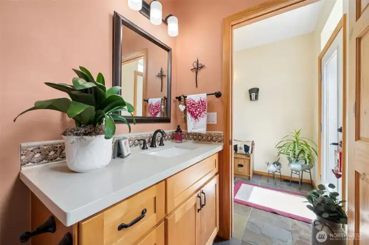 The placement of this powder room is honestly incredibly smart because it sits right off the foyer where guests can freshen up immediately upon arriving without wandering through the private areas of the home. It also works beautifully for everyday life since it is positioned perfectly for those last-minute mirror checks and quick touch-ups before heading out the door. The space itself feels far more elevated than a typical basic powder room thanks to the extra-long countertop, oversized undermount sink and warm shaker-style vanity cabinetry that continues the same rich material palette carried throughout the house.