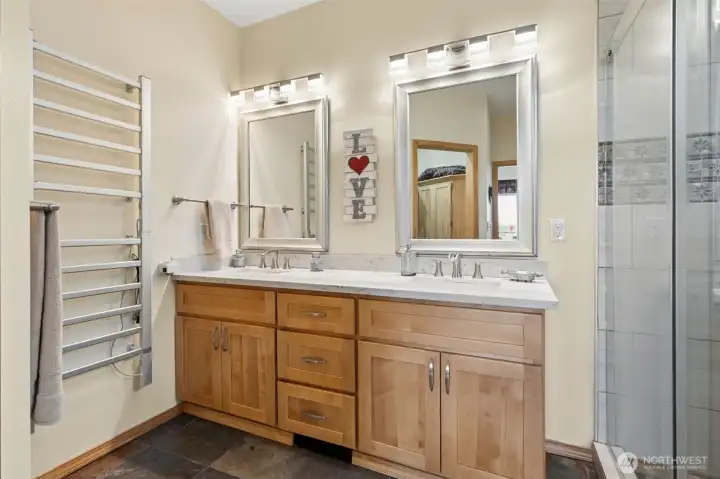 This en suite bathroom feels like exactly what you would hope to find inside a custom mountain retreat because it balances comfort, warmth and practicality without losing the polished upscale feel running throughout the home. Heated slate flooring continues into the space keeping those rich earthy textures flowing seamlessly from the primary suite, while the oversized heated towel rack adds the kind of everyday luxury that quietly spoils people wonderfully. The long double vanity offers granite counters, warm maple shaker cabinetry, framed mirrors and brushed nickel fixtures that all work together beautifully to create a timeless, grounded aesthetic with just enough lodge-inspired warmth, and over to the right, the huge glass walk-in shower starts making its appearance because apparently this bathroom intends to continue aggressively exceeding expectations. Also, there is also a dedicated toilet room tucked behind a pocket door for added privacy, plus a large walk-in closet.