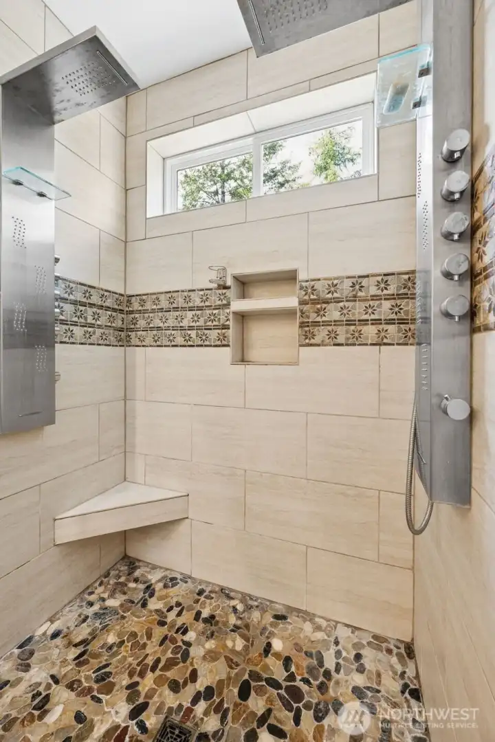Now THIS is a shower. The oversized glass walk-in design feels straight out of a luxury mountain lodge spa with enough room for two, dual shower systems and all the little thoughtful details that make the entire experience feel elevated instead of purely functional. Underfoot, the polished river rock flooring adds incredible texture and warmth while still feeling smooth and comfortable, and the tile walls are finished with decorative accent detailing that keeps the entire space visually interesting without becoming busy or trendy. Each shower station includes its own rain-style fixture and body sprays turning regular showers into something significantly more dramatic than they technically need to be, which honestly feels appropriate for this property. Built-in shelving keeps products neatly tucked away, a footrest makes shaving or rinsing easier and the long horizontal privacy window above allows fresh mountain air to flow through the space while still maintaining complete privacy.