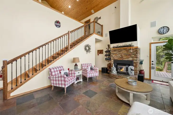 This view captures exactly why the great room feels so memorable the second you walk in because the scale, textures and architectural details all work together without trying too hard to impress you. The dramatic staircase climbs upward beneath soaring vaulted wood-plank ceilings creating this incredible lodge-inspired focal point, while the combination of black iron railing details and rich wood handrails gives the whole space a more elevated custom-home feel compared to traditional all-wood stair systems. Warm cream walls keep the room feeling bright and inviting despite the dramatic ceiling height, and the entire space stays visually connected to the heated slate flooring and mountain views beyond. Underneath the staircase, additional storage is tucked neatly away while the floorplan itself loops naturally through the primary suite hallway and back toward the foyer creating a circular main-floor layout that feels wonderfully fluid and easy for both everyday living and entertaining.